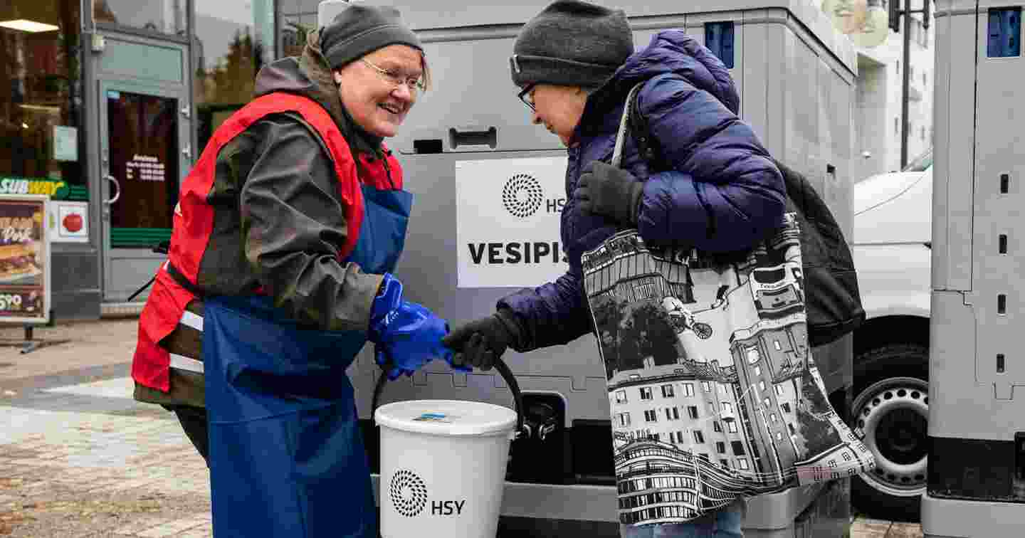 Hymyilevä nainen Punaisen Ristin vapaaehtoisliiveissä ojentaa kannellista ämpäriä naiselle, jonka vedenjakelu on katkennut. Punainen Risti suorittaa vedenjakelua kotimaan kriisitilanteissa.